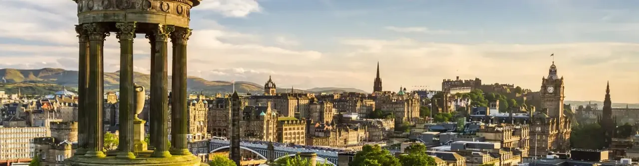 Classical columns frame a panoramic view of Edinburgh's historic cityscape with castle, spires, and surrounding hills under cloudy sky.