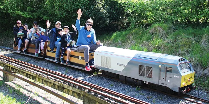 An operator and children riding in open carriages pulled by a miniature diesel locomotive through wooded countryside.