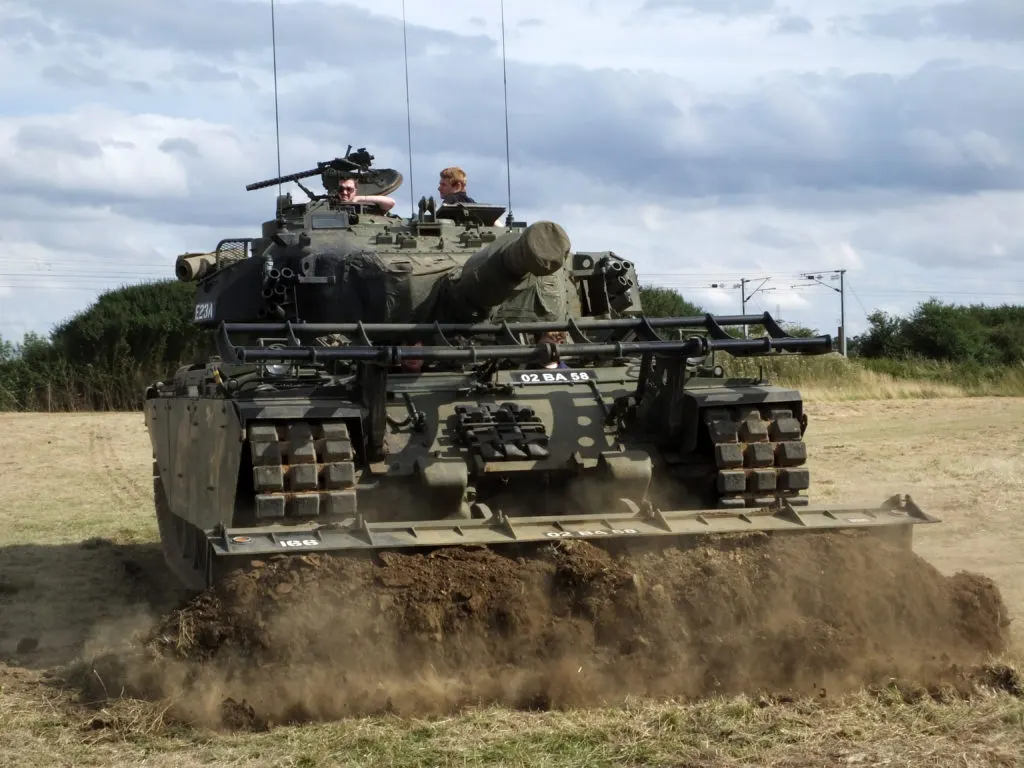 Military tank with two operators on board driving through muddy terrain, creating a dust cloud.