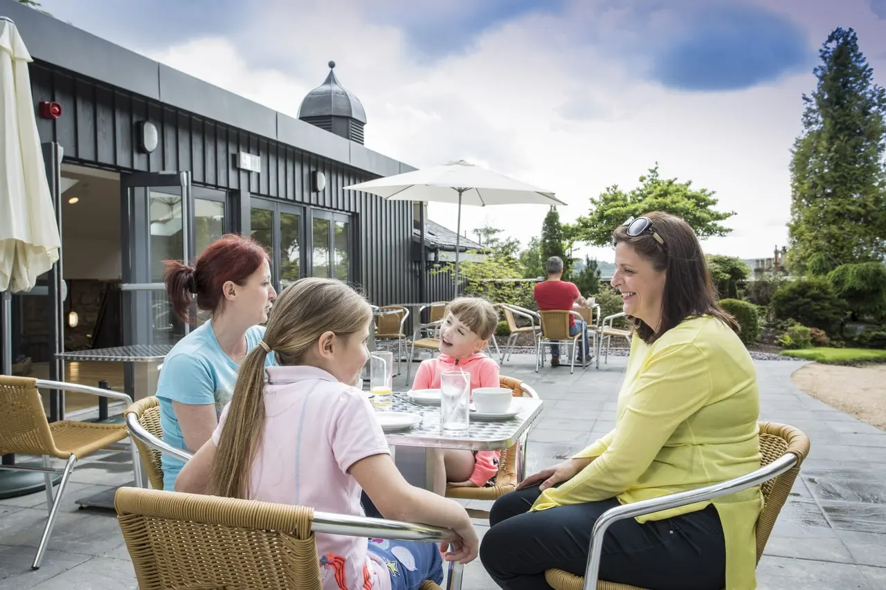 Adults and children dining and socialising at outdoor terrace seating with umbrellas, modern building and landscaped gardens in background.