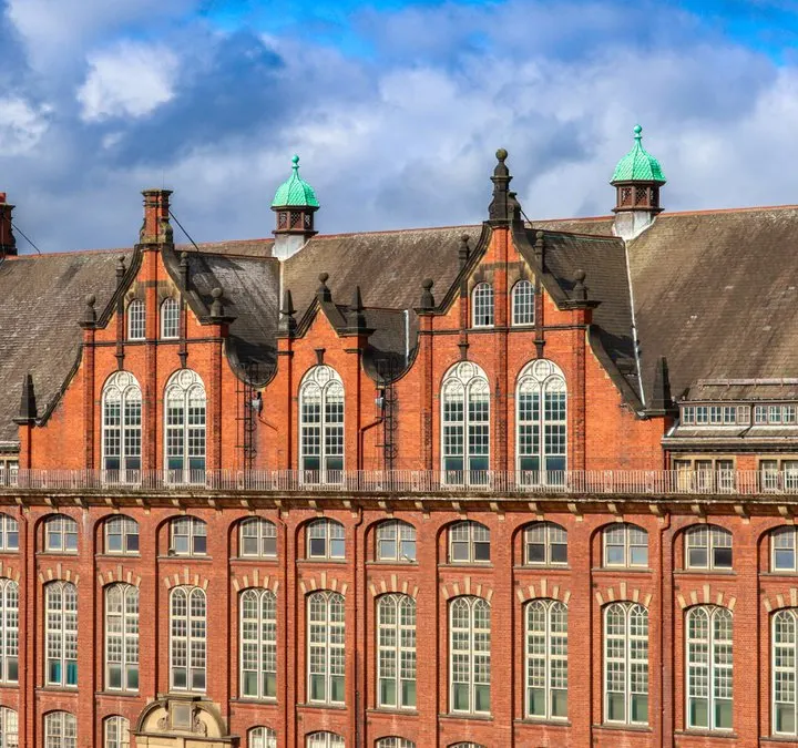A red-brick Victorian building with ornate architecture, featuring green copper domed turrets and rows of tall arched windows against a blue cloudy sky.