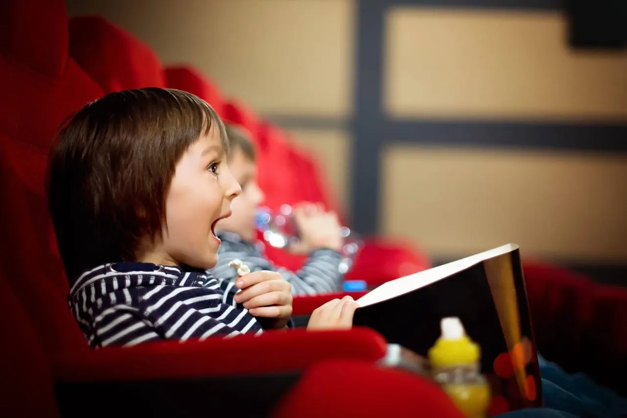 Young child seated in a red cinema chair holding popcorn and watching a film.