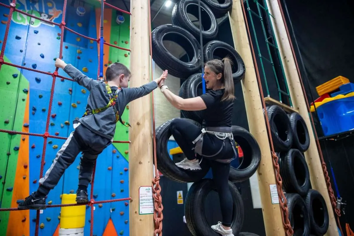 Child climbing colourful climbing wall with handholds whilst adult instructor high-fives from tyre obstacle course nearby.