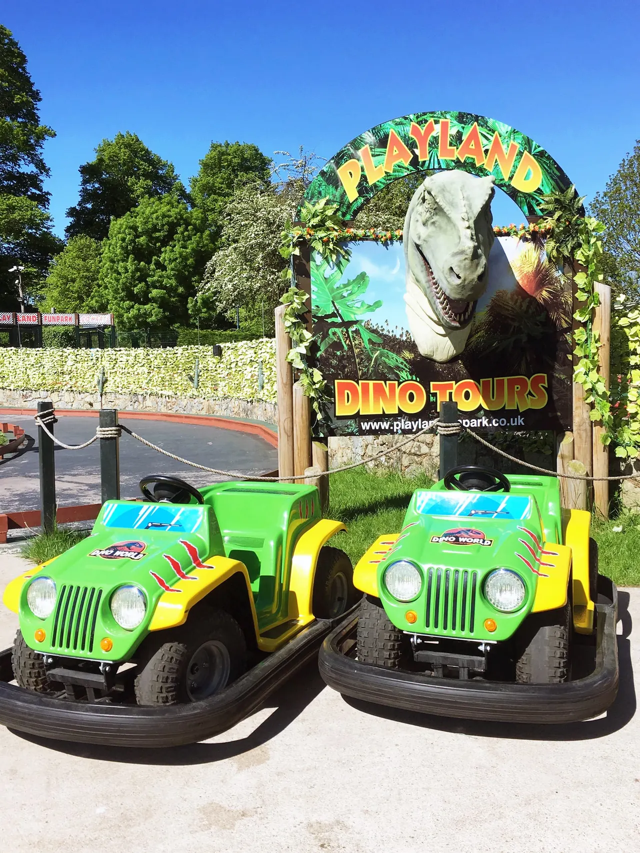 Bright green and yellow dinosaur-themed ride cars parked beneath an arch sign for dino tours at an outdoor amusement park.