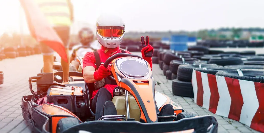 Go-kart driver in red and orange kart making a peace sign on an outdoor racing track with tyres lining the course.