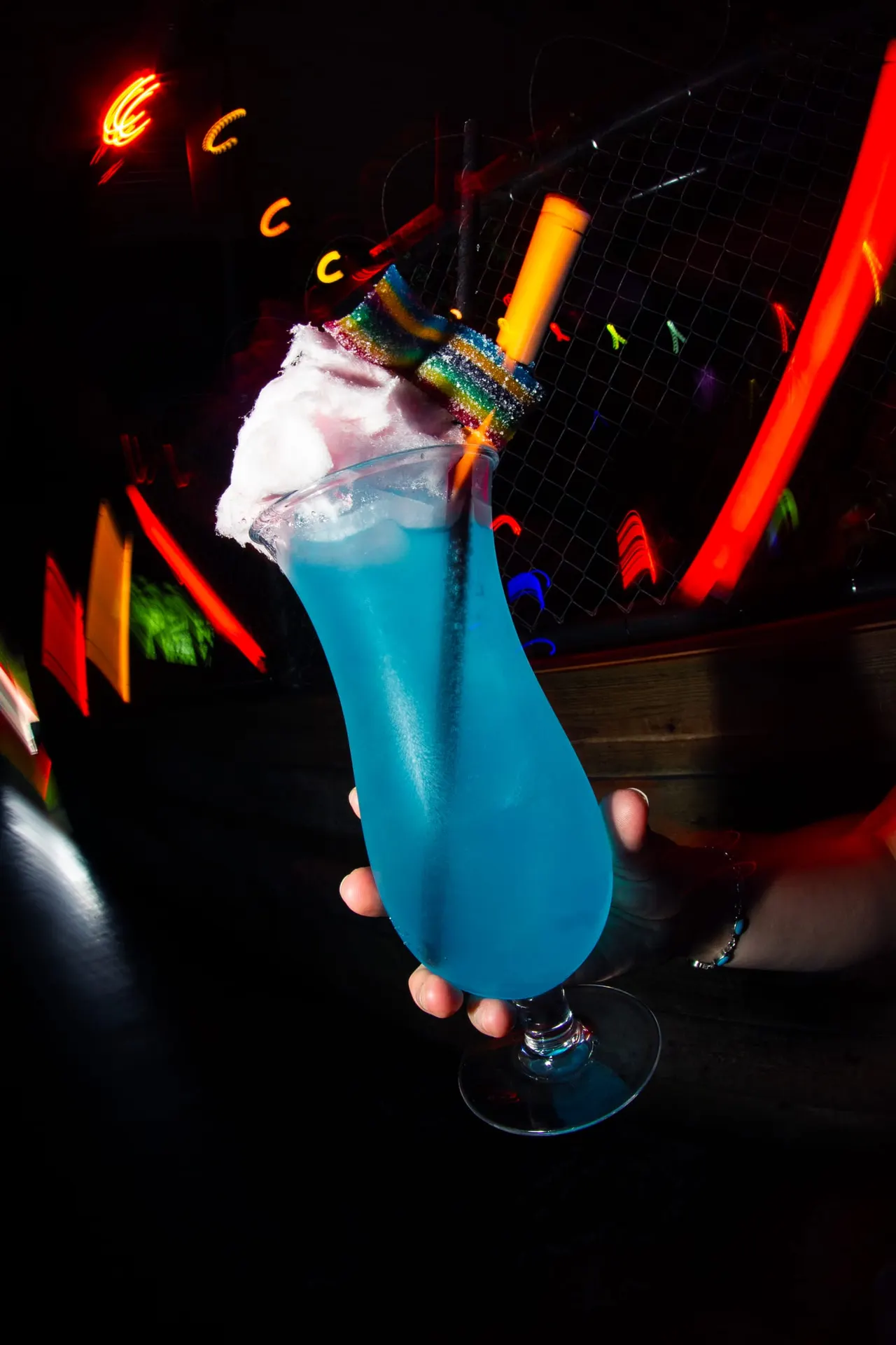 Hand holding a bright blue cocktail with cotton candy and a yellow straw, surrounded by neon signs in a dark venue.
