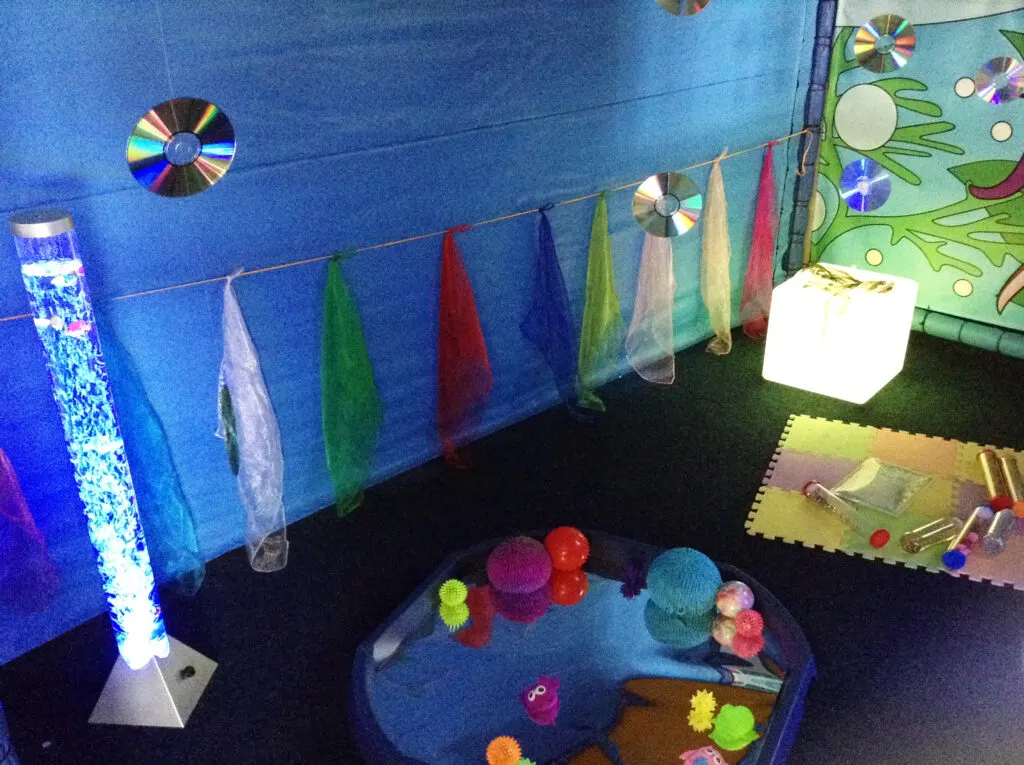 Sensory play area with coloured fabrics strung across blue walls, glowing light tubes, and textured balls and toys on a platform.