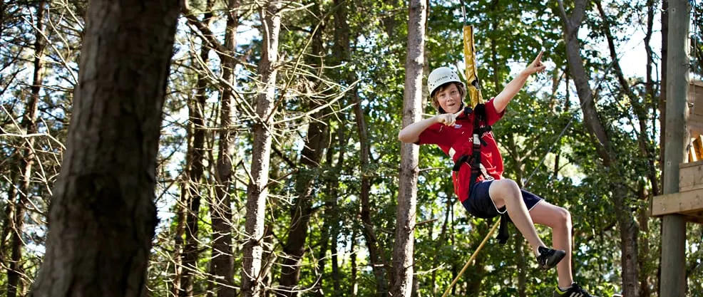 Child in red shirt zip-lining through a forested canopy course amongst tall trees.