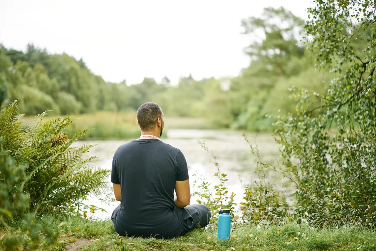 Person sitting on the ground beside a river surrounded by lush woodland, contemplating the water.