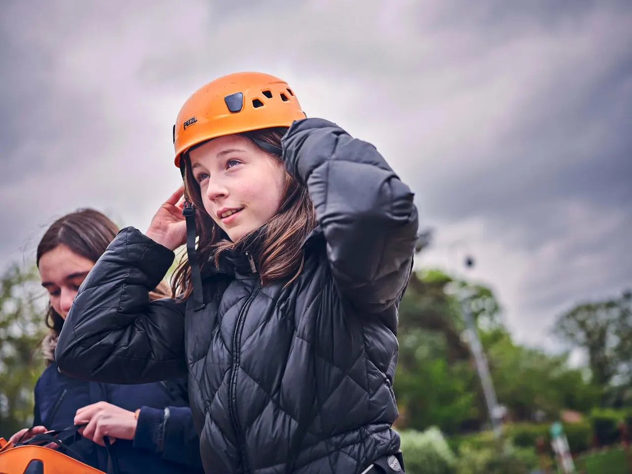 A young person wearing an orange safety helmet and black padded jacket fastening their helmet strap, with another child beside them outdoors.