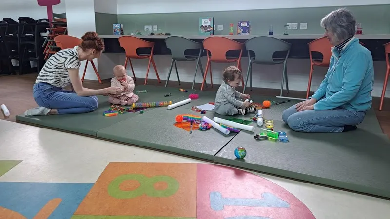 Two adults and two toddlers playing with colourful toys on a padded floor in an indoor activity space.