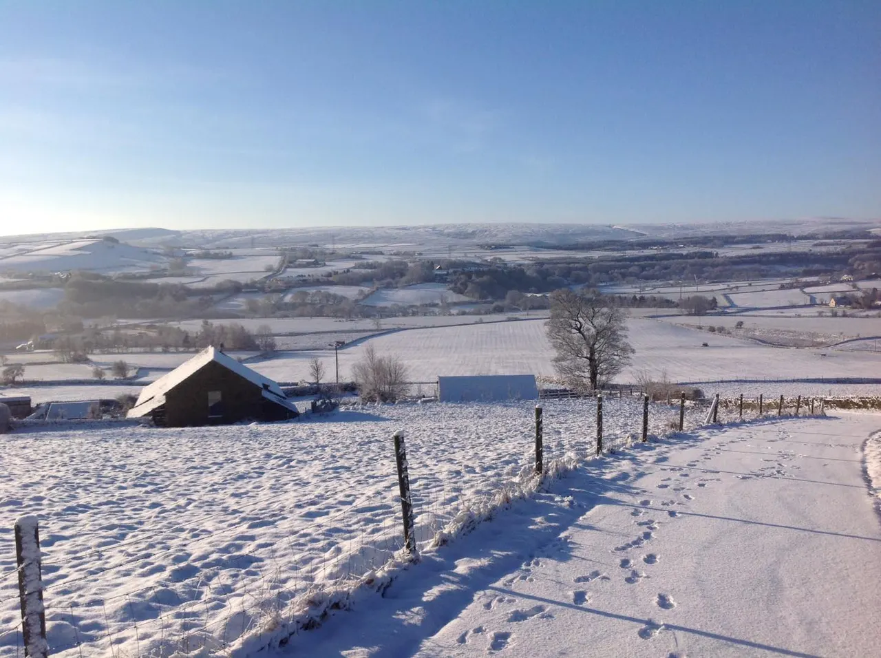 Snowy farmland with barns and fences in the foreground, rolling countryside and fields stretching to the horizon under clear blue sky.