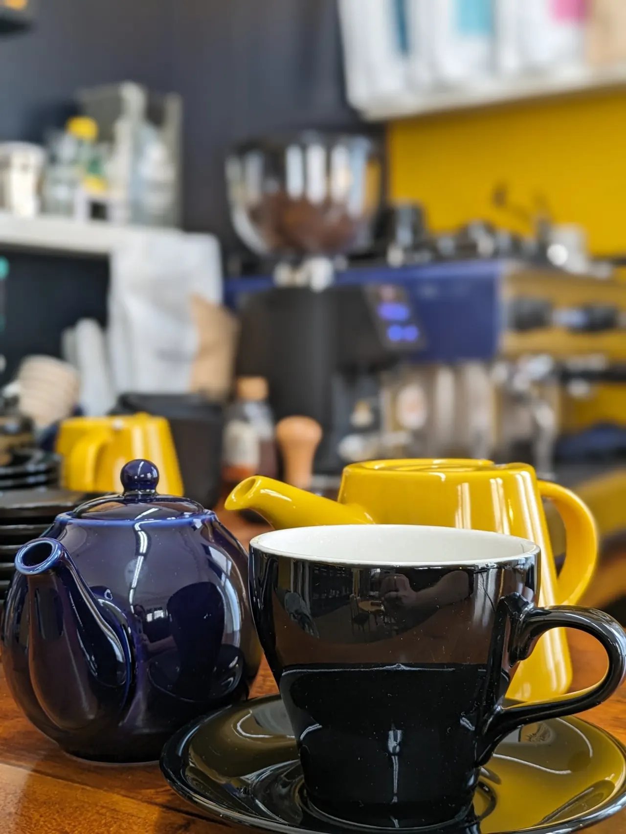 Colourful ceramic tableware including a dark blue teapot, black cup and saucer, and yellow jug displayed on a wooden counter in a bright leisure venue.