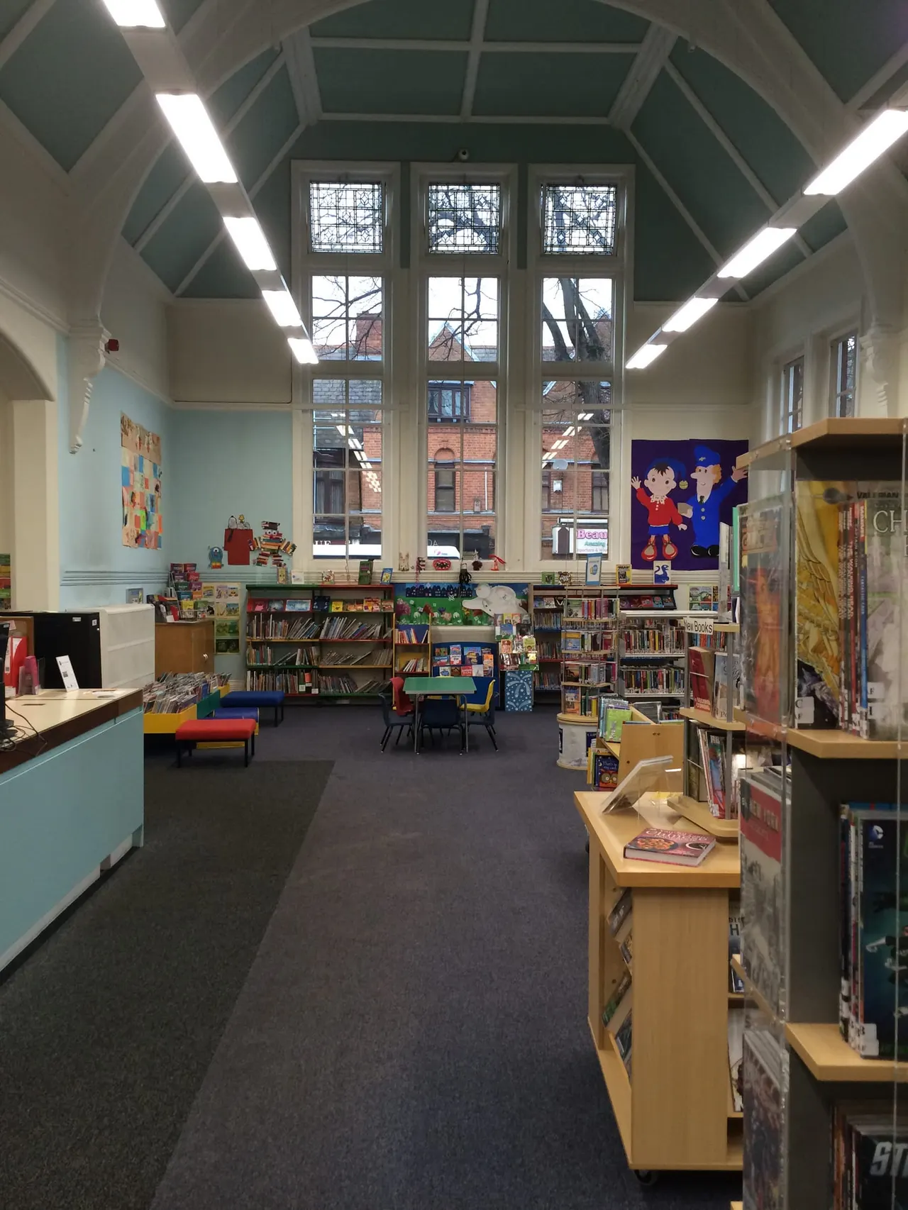 Bright children's library interior with bookshelves, colourful seating, artwork on walls and tall windows overlooking a street.