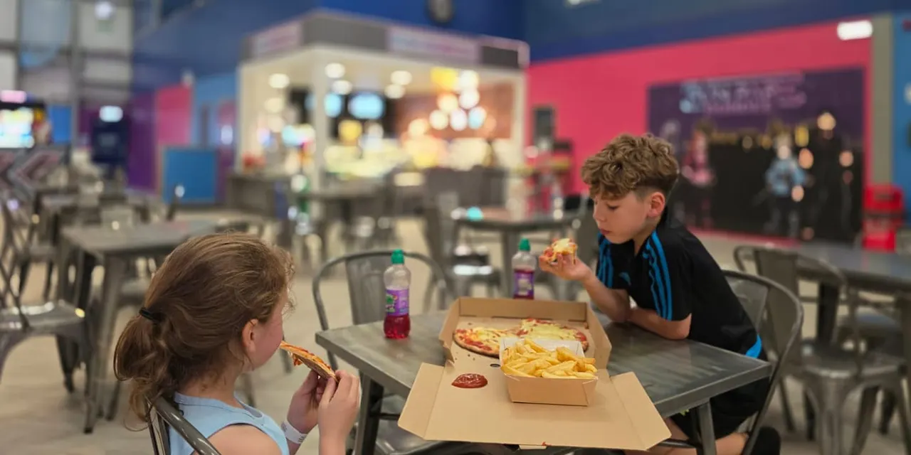 Two children eating pizza and chips at a table in a colourful indoor leisure venue with arcade games and a food counter.