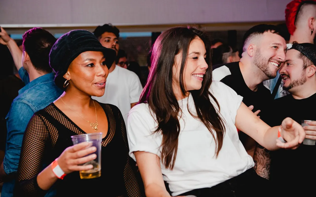 Young people enjoying drinks and socialising together on a lively dancefloor with purple lighting.