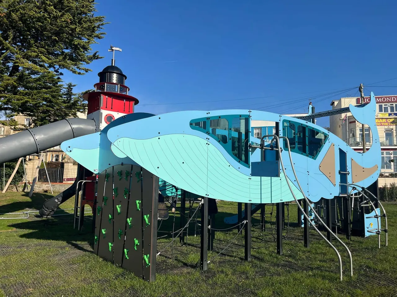 A blue whale-themed play structure with climbing wall and slide on grass, with a red and white lighthouse in the background.