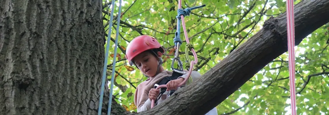 Child in red helmet on tree-top course secured with ropes and safety equipment among green foliage.