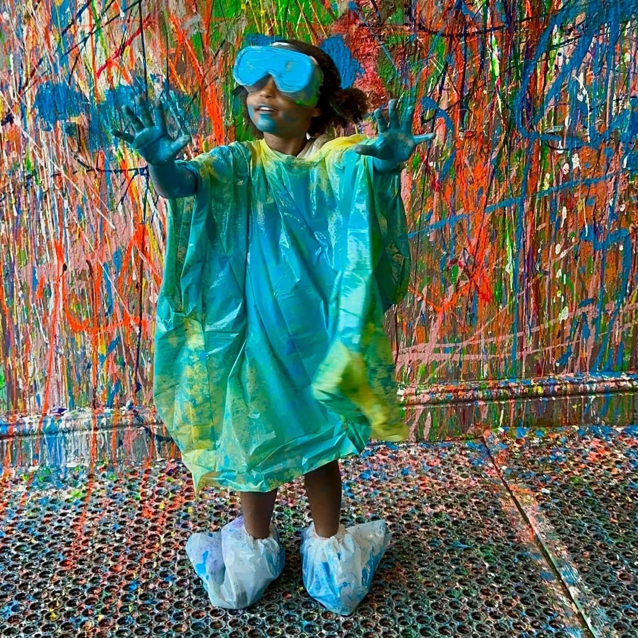 Child in protective gear posing with arms outstretched inside a colorful paint-splattered interactive art room.