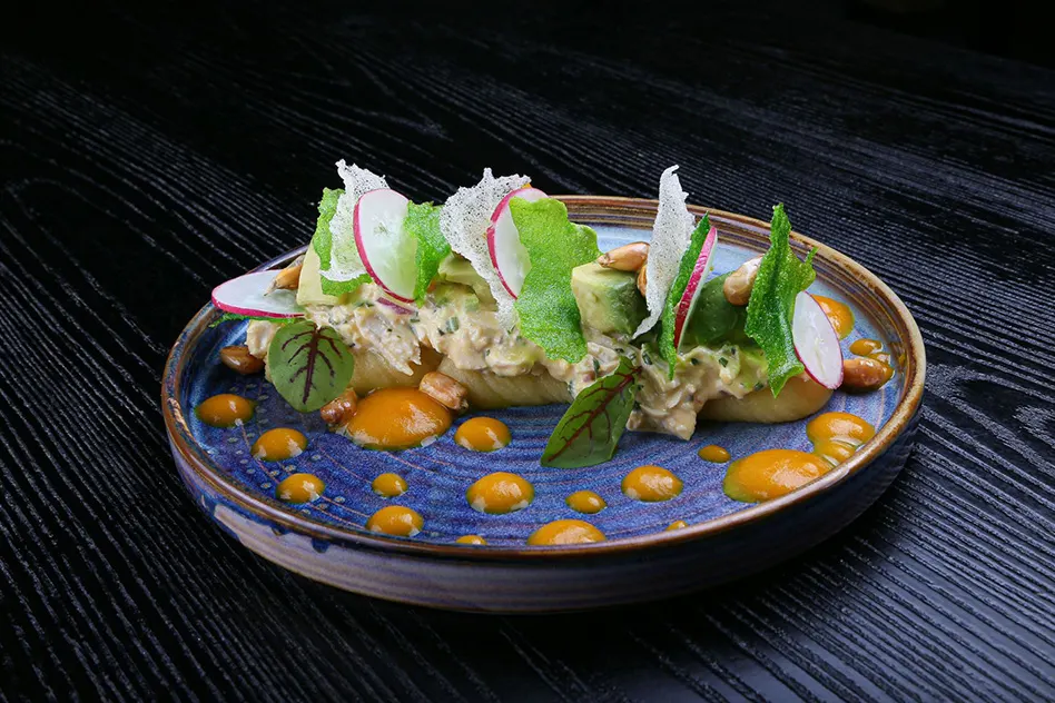 Artfully plated seafood dish with radish slices, green herbs, and golden roe on a blue-glazed plate against dark wood.