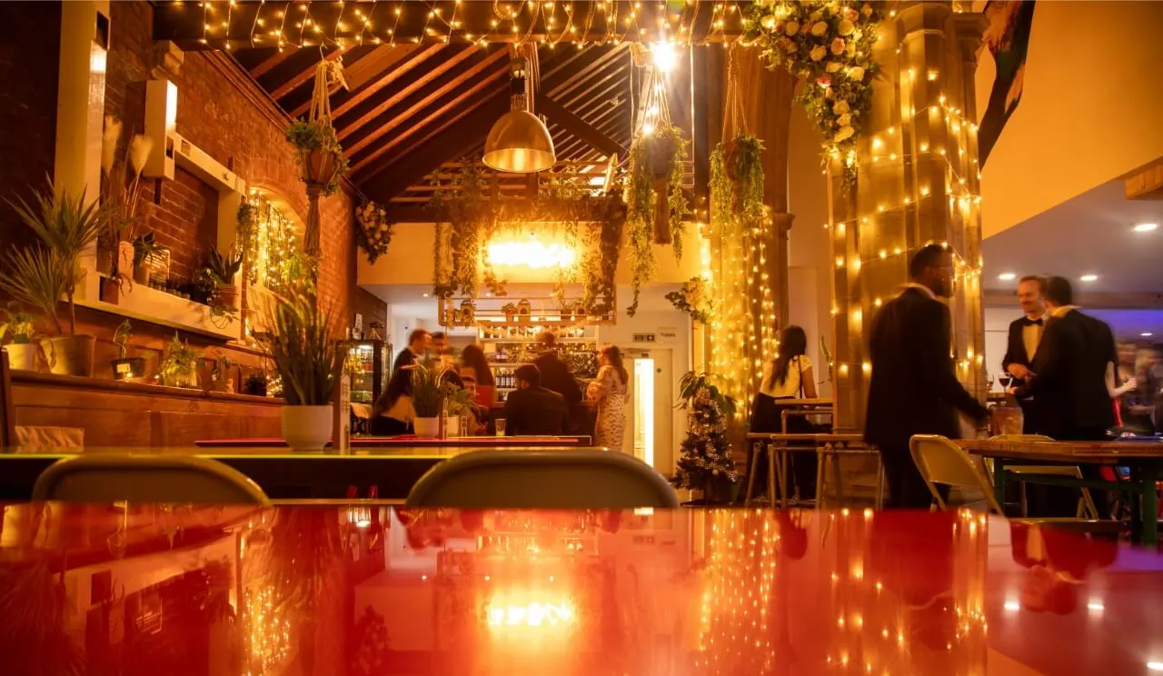 Interior of an upmarket bar decorated with warm lighting, hanging plants, and string lights, with staff and customers visible at the counter and throughout the space.