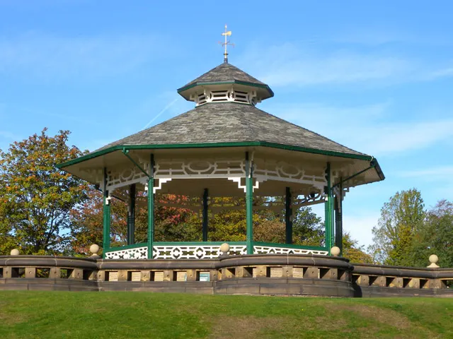 Victorian bandstand with green-painted woodwork and cross-topped roof in parkland setting with autumn trees.