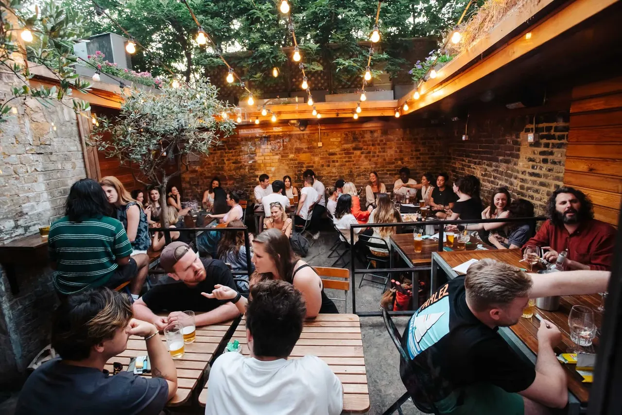 Busy outdoor courtyard with brick walls, wooden beams, and hanging lights, where a large group of people sit at tables drinking and socialising.