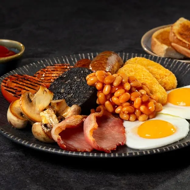 A full cooked breakfast plated on dark ceramic with bacon, sausages, mushrooms, baked beans, eggs, toast and grilled tomato.