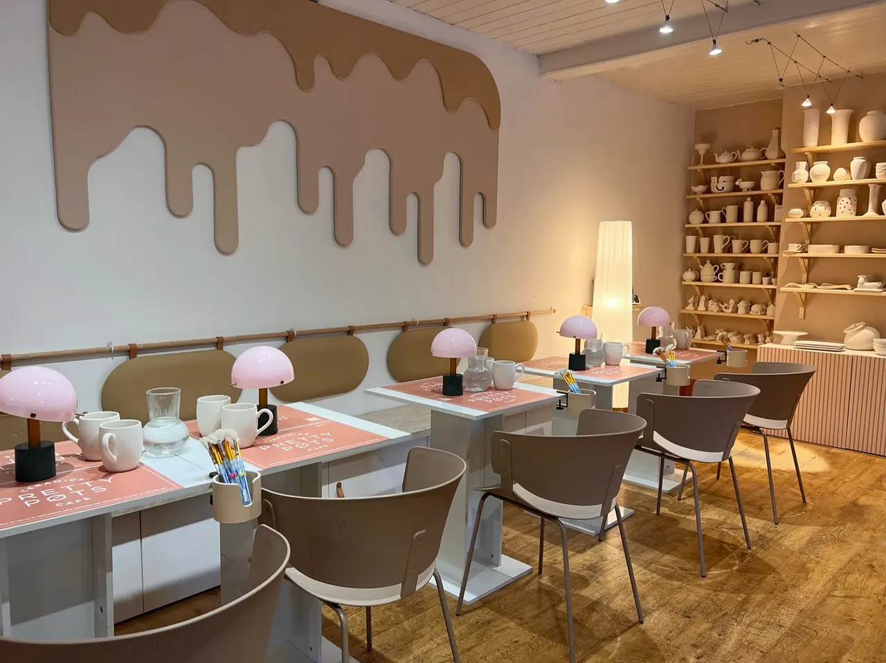Modern pottery studio with long work tables, pink dome lamps, ceramic supplies, and shelving displaying finished pieces.