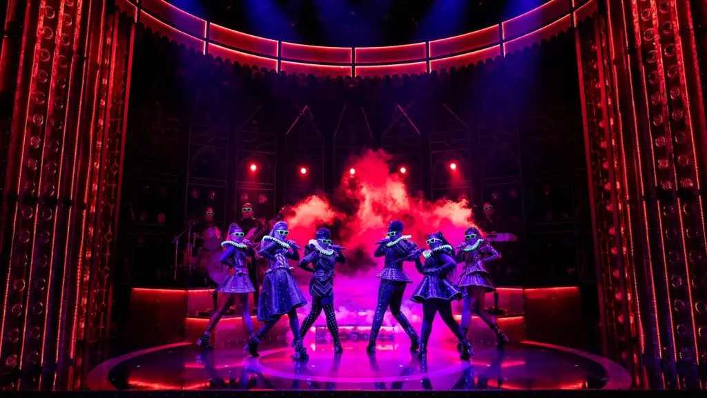 Six performers in coordinated costumes dance on stage beneath red neon framing and pink smoke effects.