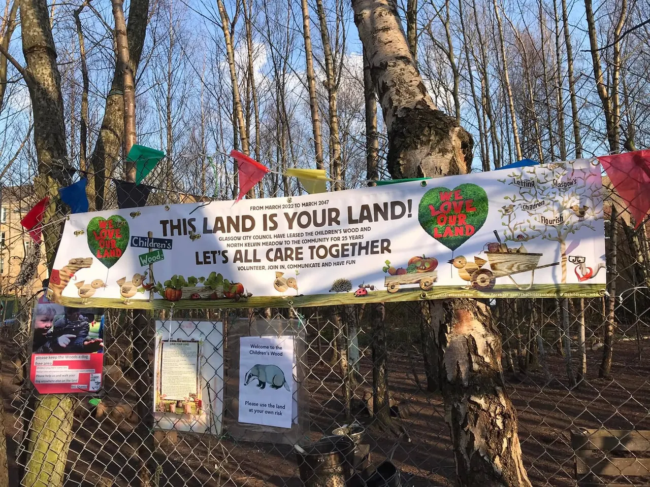 Welcome banner and information signs at the entrance to a wooded community space, with bare trees and colourful bunting overhead.