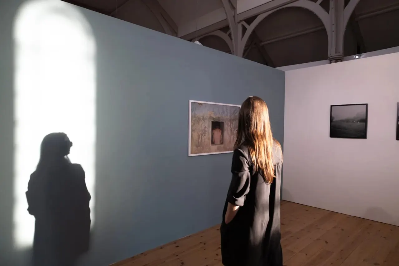 Woman viewing framed photographs in an art gallery with blue and pink walls and industrial ceiling.