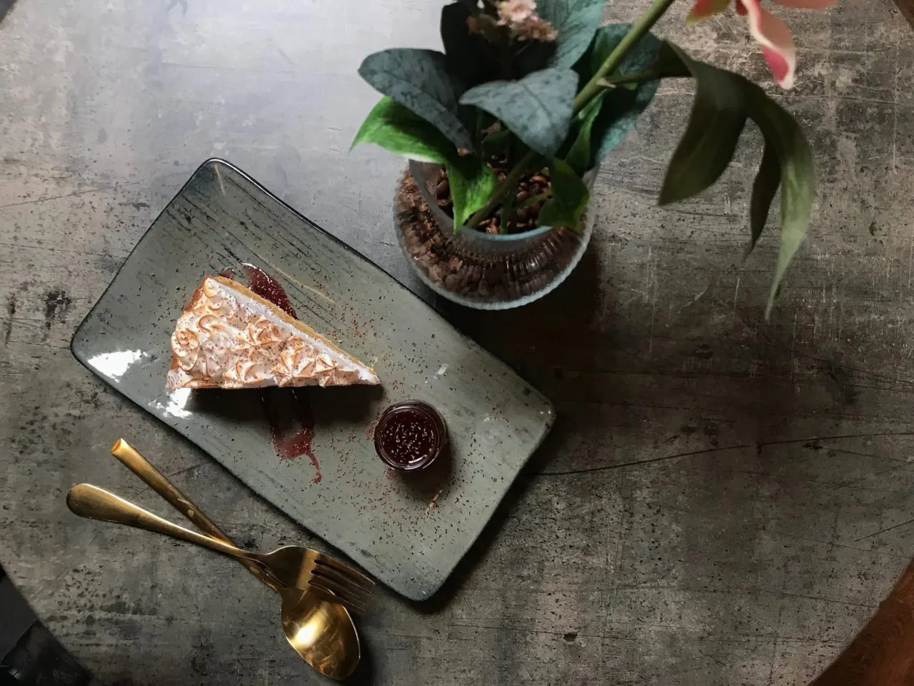Slice of layered cake on a grey plate with jam and gold cutlery, beside a potted plant on a concrete table.