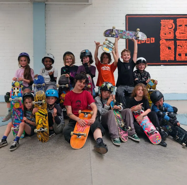 Group of young skateboarders posing together indoors holding their skateboards, wearing helmets and protective gear.