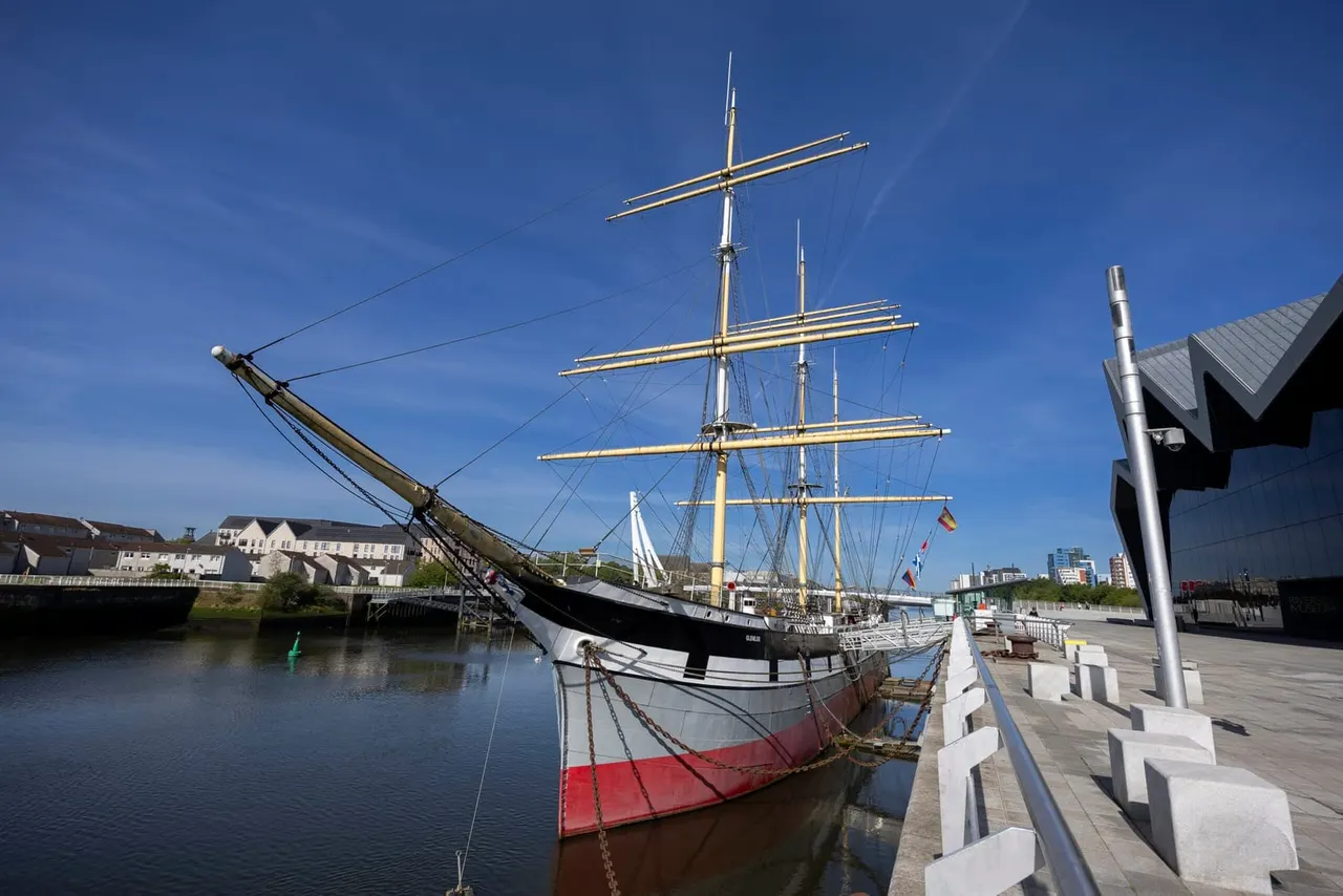 Tall ship with golden yards and rigging moored at a waterside promenade on a clear day.