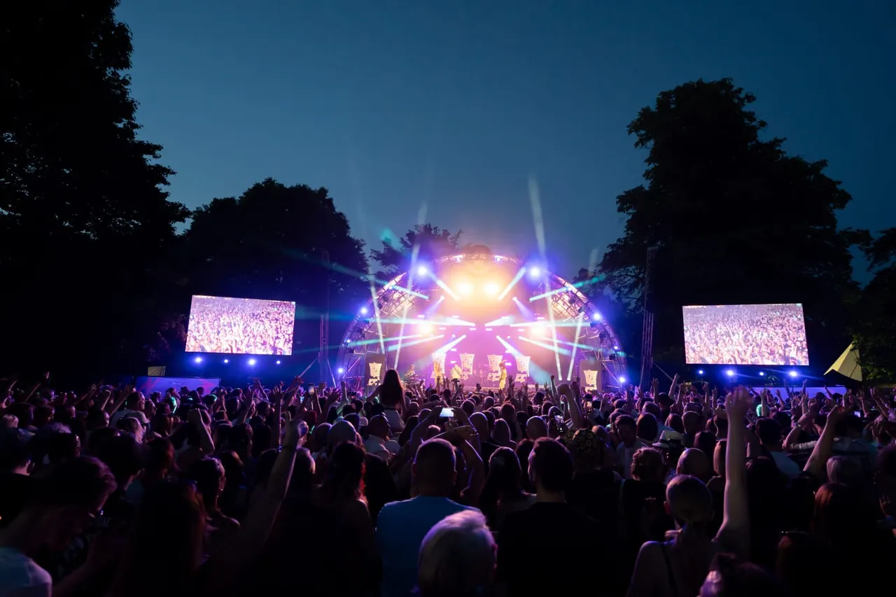 Large outdoor festival crowd dancing in front of illuminated stage with screens at night amongst trees.