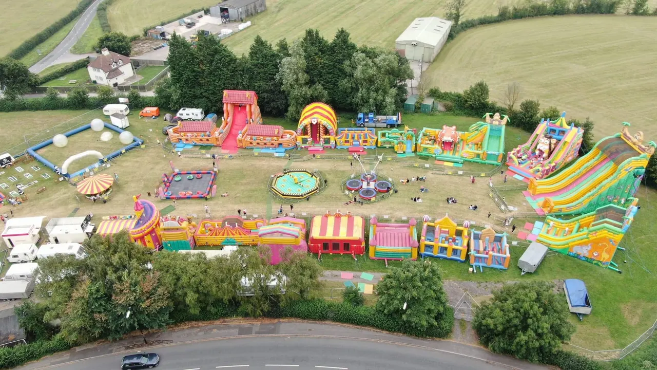 Aerial view of an inflatable theme park in a rural field with colourful rides, slides, and attractions set up on grass.