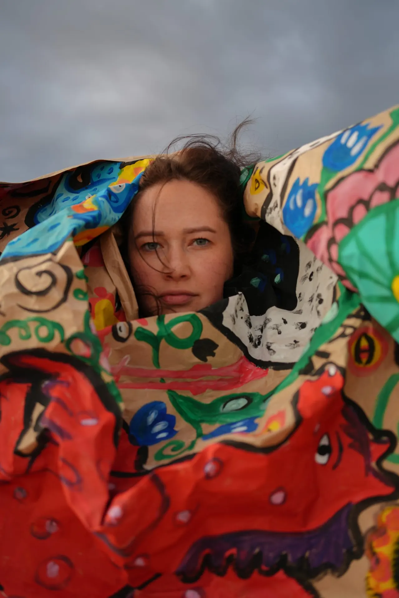 Person holding a large colorful kite with artistic designs against a grey sky.