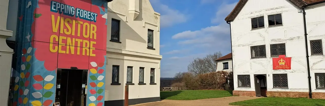 Historic buildings with signage for Epping Forest Visitor Centre, set in countryside with grassy grounds under blue sky.