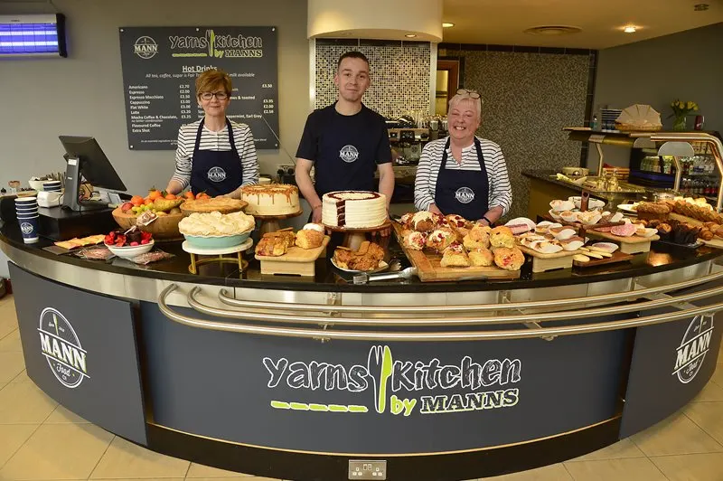 Three staff members stand behind a curved counter displaying various baked goods, cakes and pastries at Yams Kitchen.