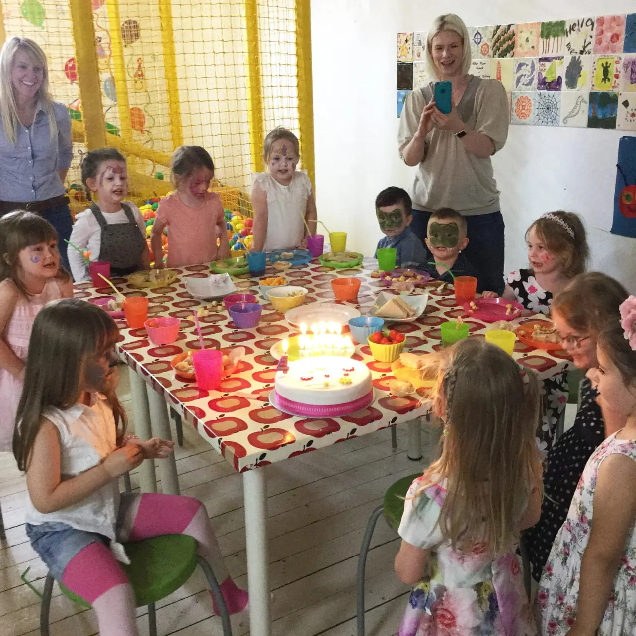 Children gathered around a table with a birthday cake during an indoor party, some wearing face paint.