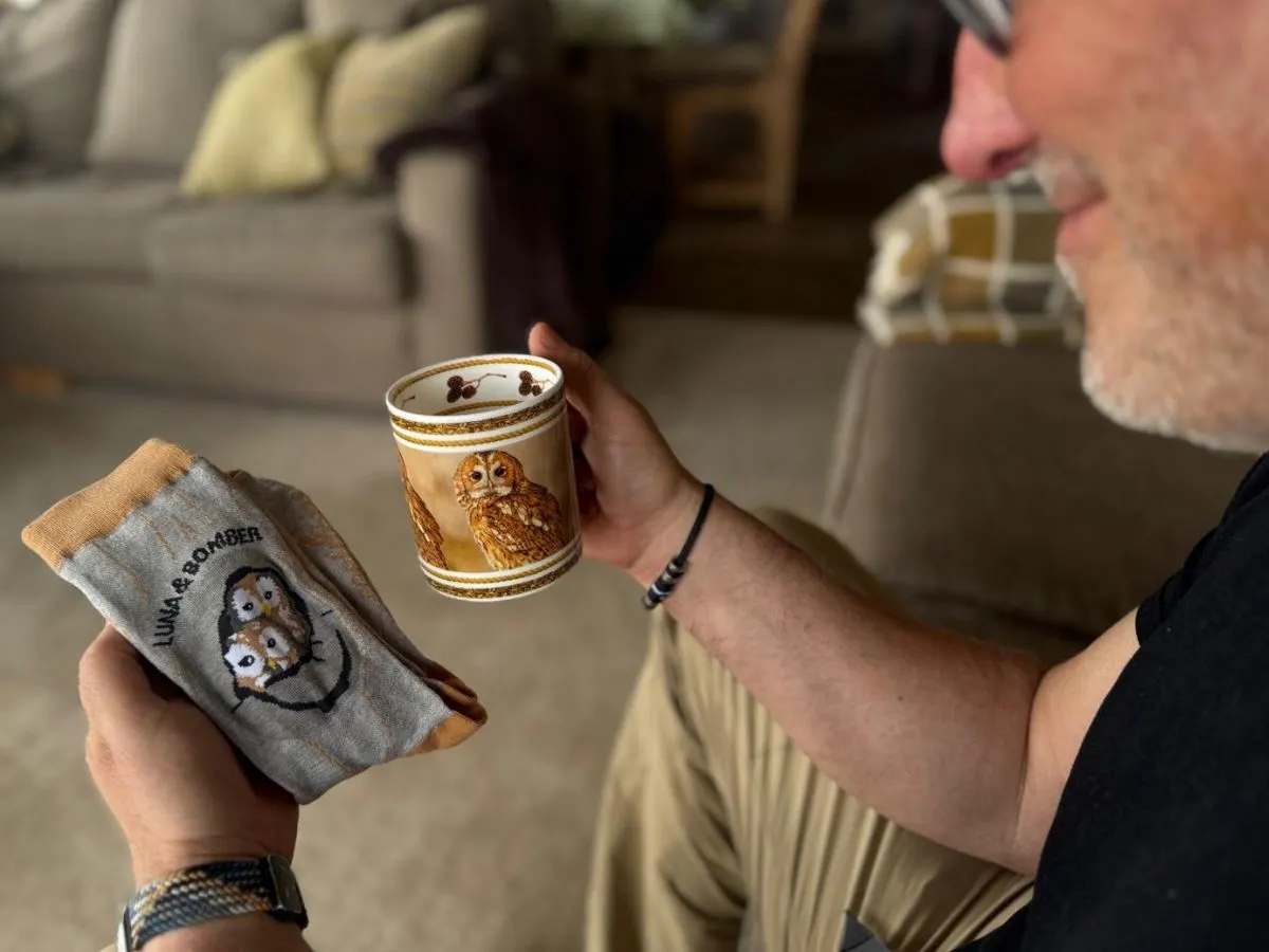 A man holding an owl-themed mug and cloth merchandise at an indoor venue.