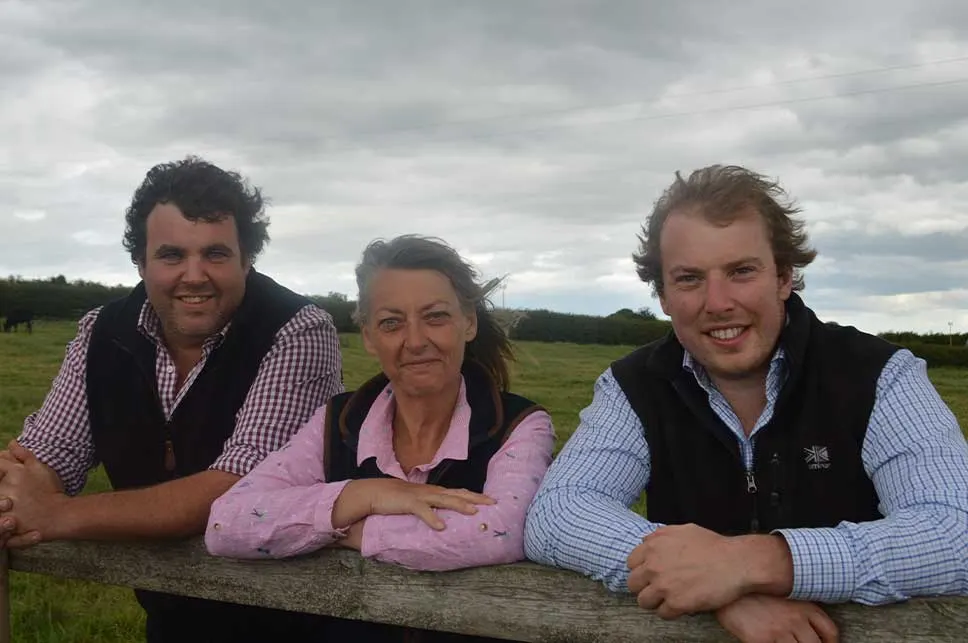 Three people leaning on a wooden fence in a green pasture with cloudy sky behind them.