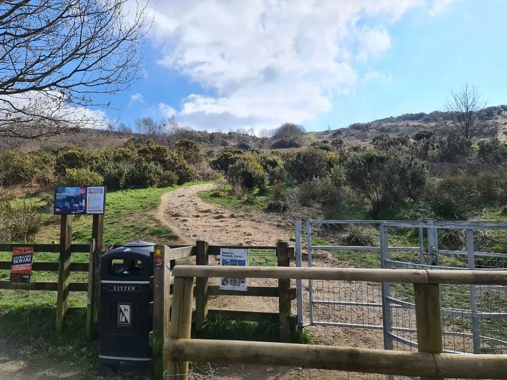 Entrance gate to a woodland walking trail with information boards, a litter bin, and a dirt path leading through trees and rolling hillside.