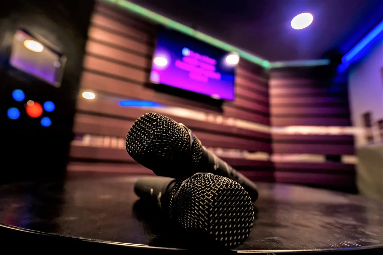 Two microphones on a stage with neon-lit karaoke screens and colourful lighting in the background.