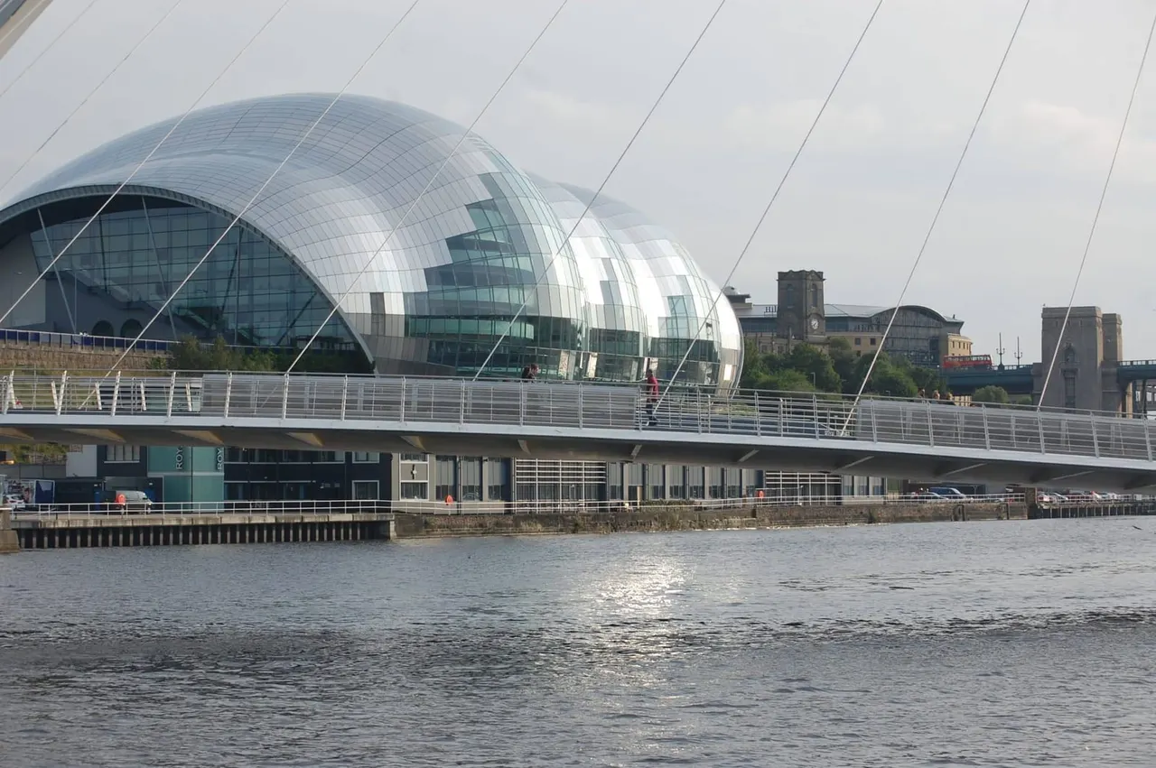 Modern curved glass building on waterfront with pedestrian bridge crossing river.