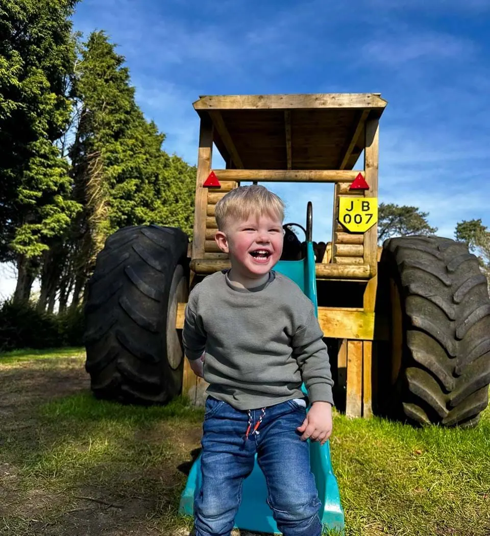 Young child laughing while sitting in a toy tractor with large tyres in a grassy outdoor setting.