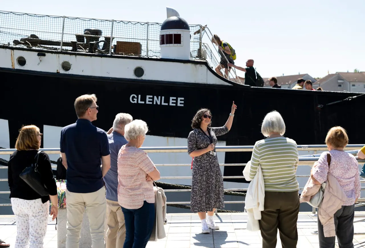 A tour guide addresses visitors boarding the Tall Ship Glenlee at a waterfront dock.