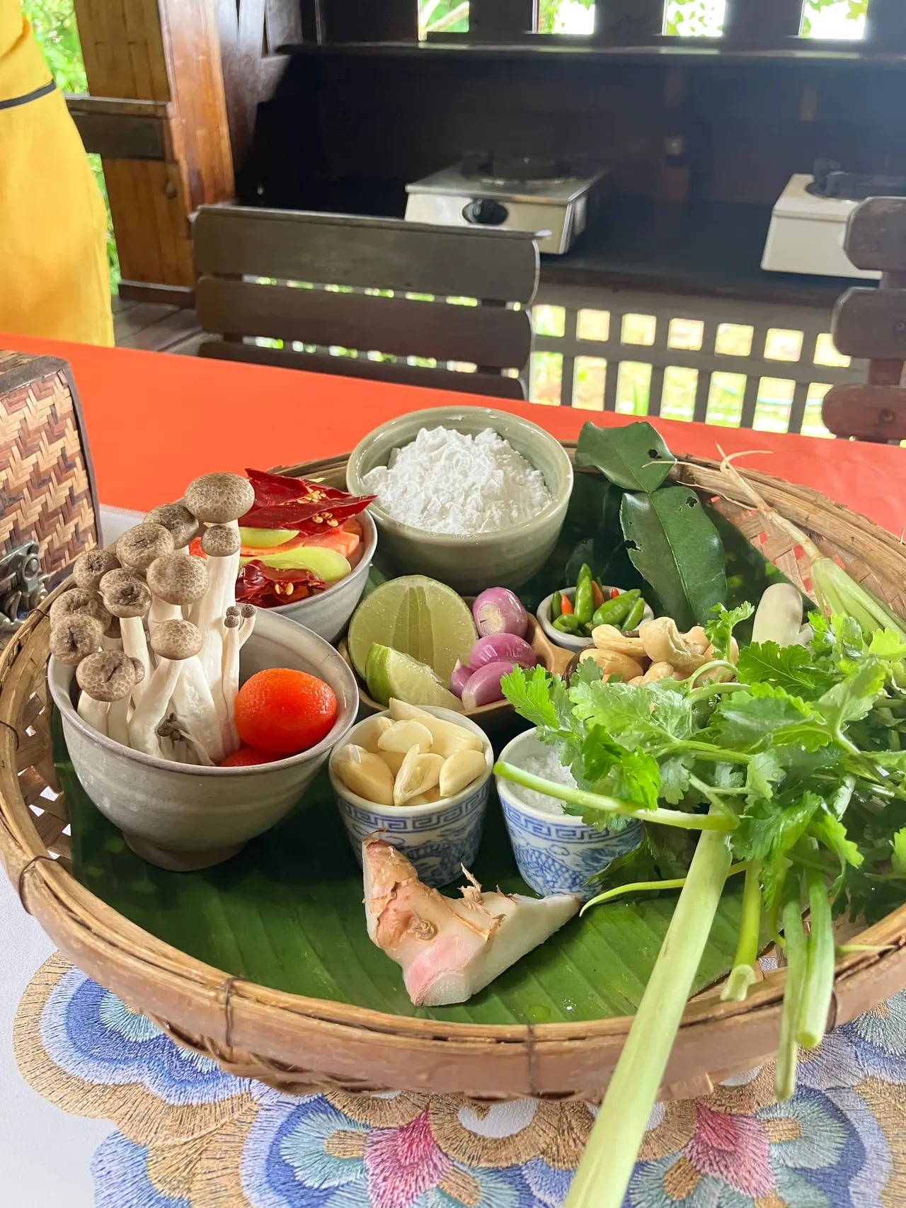 Arranged ingredients for Asian cooking displayed on a wooden tray with banana leaf, including mushrooms, chillies, lime, onions, garlic, herbs and spices.