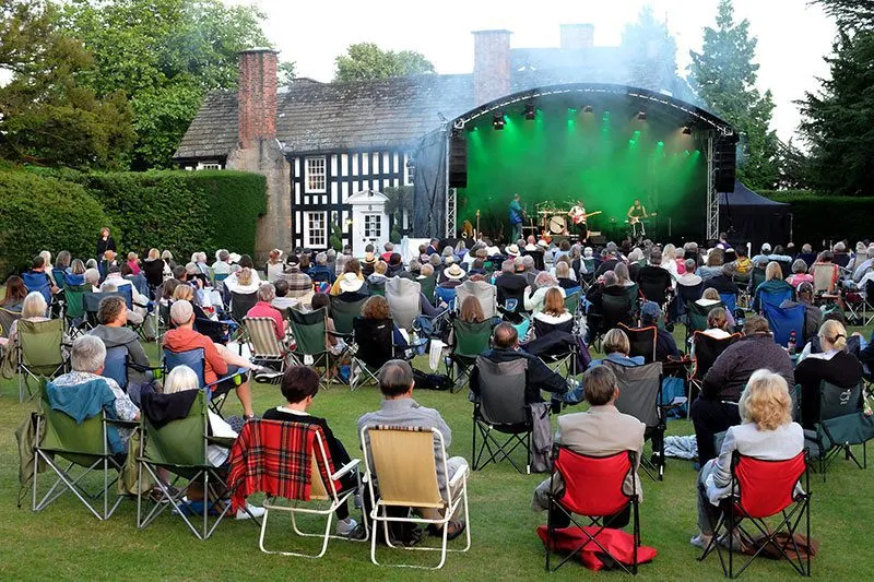 Outdoor concert with crowd seated on lawn facing illuminated stage near historic hall and gardens.